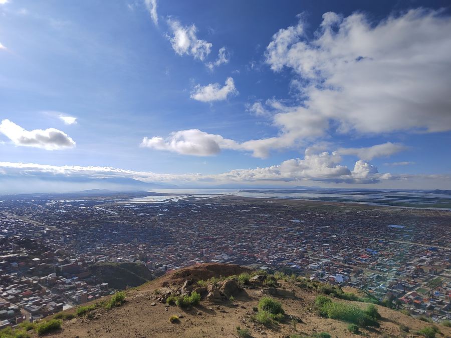 Vista desde un cerro sobre la ciudad de Oruro, extendida en la llanura del altiplano, con el lago Uru Uru al fondo bajo un cielo con nubes.