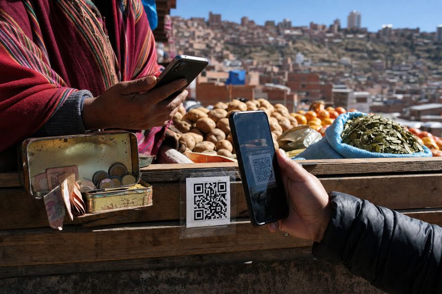 Puesto de mercado en La Paz: un cartel plastificado con un código QR pegado al mostrador junto a una lata con monedas y billetes, mientras dos manos con celulares hacen el pago.