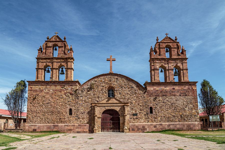 Fachada de piedra rojiza del templo de Laja, con dos torres de campanario y una cruz sobre el arco central, bajo un cielo azul.