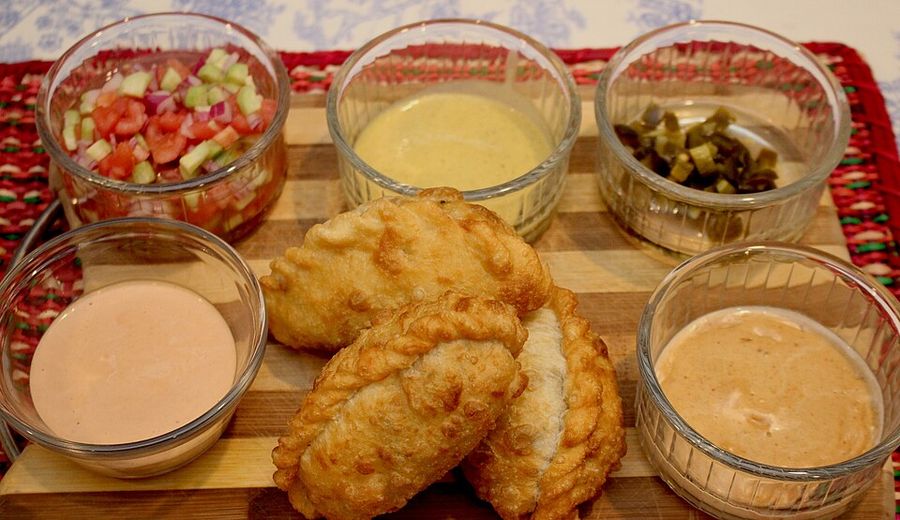 Tucumanas fritas sobre una tabla, rodeadas de cinco cuencos con salsas: llajua de tomate, salsa de maní, escabeche de verduras y dos salsas cremosas.